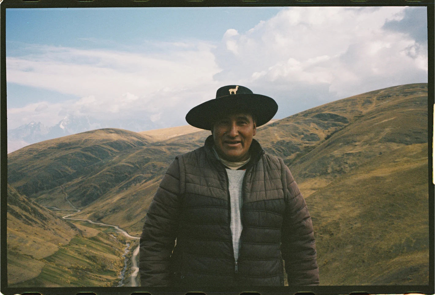 Felipe, an alpaquero, standing in the Andes at sunrise wearing a black hat with an alpaca emblem and quilted jacket