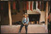 Quechua woman seated on a stone step outside a workshop with colorful handwoven textiles hanging overhead