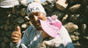 Felipe smiling and giving a thumbs up while holding a baby in a pink hat in front of a stone wall, wearing a white beanie labeled PAKA