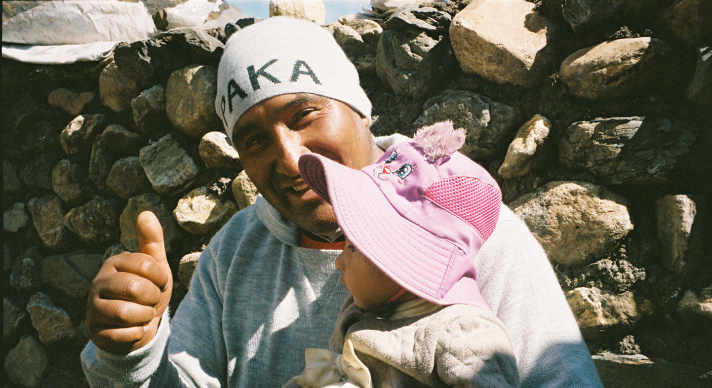 Felipe smiling and giving a thumbs up while holding a baby in a pink hat in front of a stone wall, wearing a white beanie labeled PAKA