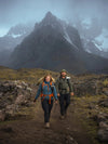 Two hikers walking on a rocky alpine trail wearing fleece layers and backpacks with dramatic misty snow-capped peaks behind them