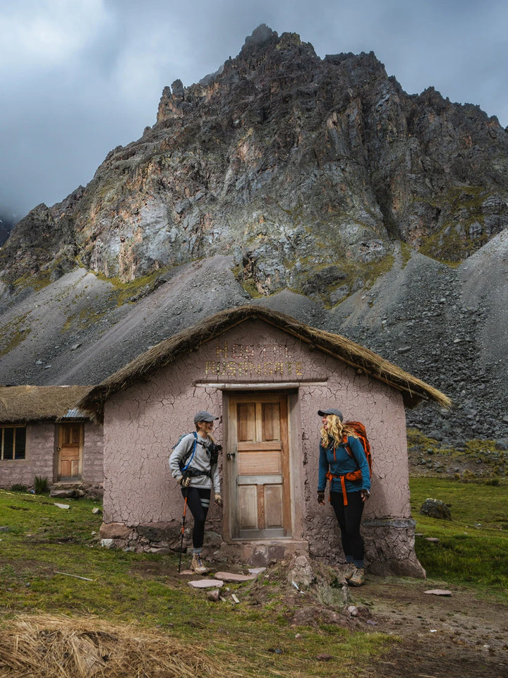 Two hikers standing outside small stone huts at the base of jagged mountain peaks in Peru
