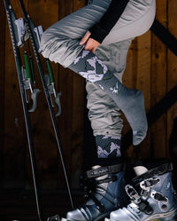 Side view of Snow Sebastian indigo ski socks on a model showing leg compression and cushioned foot with ski boots and wooden cabin background
