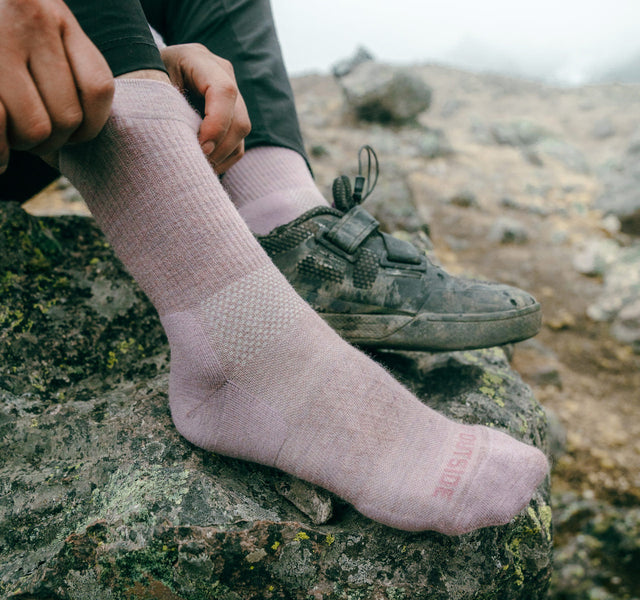 Side view of mauve performance crew sock on a hiker showing ribbed calf for grip and cushioned heel on rocky trail background