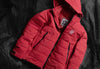 Angled flat-lay of red Huascarán puffer jacket showcasing hood, zipper, zip pockets, and PAKA logo on black net background