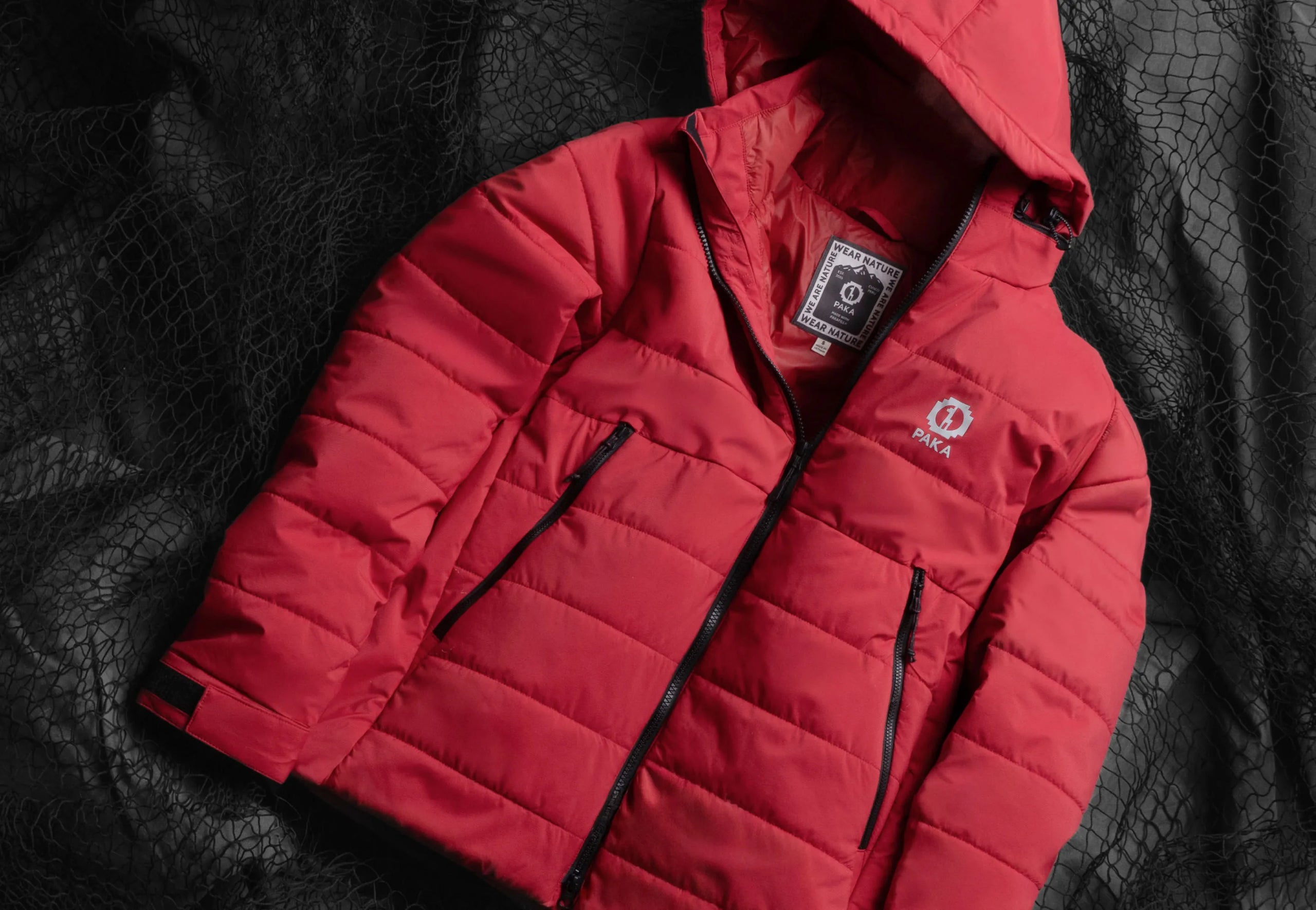 Angled flat-lay of red Huascarán puffer jacket showcasing hood, zipper, zip pockets, and PAKA logo on black net background