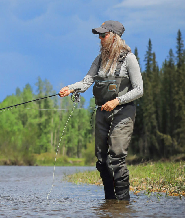 A blond woman fishing in a river in her grey alpaca baselayer