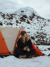 Hiker sitting at the entrance of an orange tent on snow with a snow-covered mountain in the background