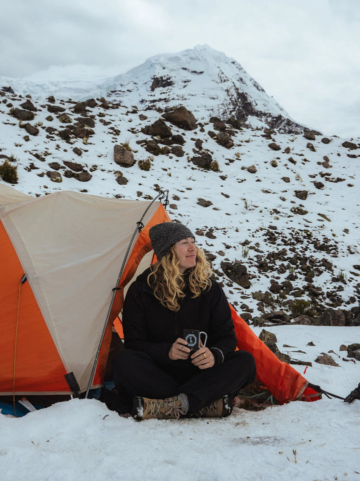 Hiker sitting at the entrance of an orange tent on snow with a snow-covered mountain in the background