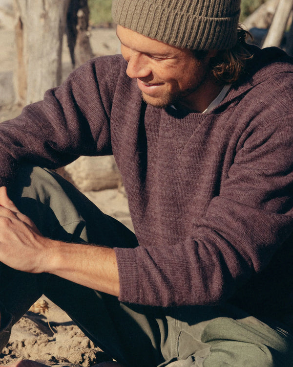 Three-quarter view of The Hoodie alpaca hoodie in raisin showing hood, relaxed unisex fit, and knit texture on a sandy beach