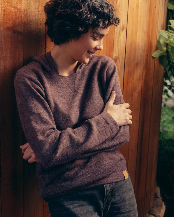 Three-quarter view of a model wearing The Hoodie alpaca knit hoodie in raisin, showing soft, relaxed unisex fit against a wooden fence background