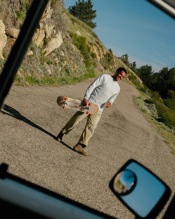 Man wearing light grey alpaca crewneck sweater in a relaxed fit walking on a sunlit country road carrying a skateboard