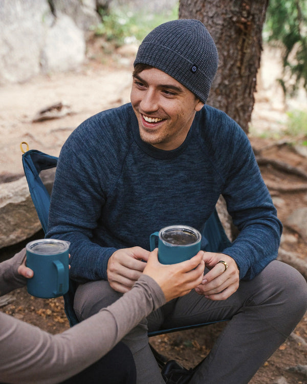 Man wearing a navy alpaca crewneck sweater in regular fit smiling and sharing a mug while seated at a campsite