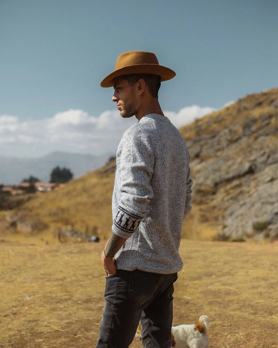 Man wearing a brown hat and gray sweater standing in a field with mountains in the background