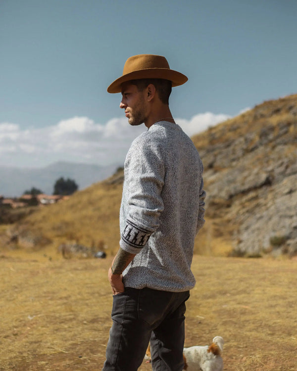 Man wearing a brown hat and gray sweater standing in a field with mountains in the background