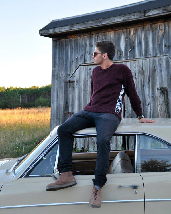 Man wearing burgundy The Cusco sweater with white alpaca jacquard side panel sitting on a vintage beige car by a wooden barn