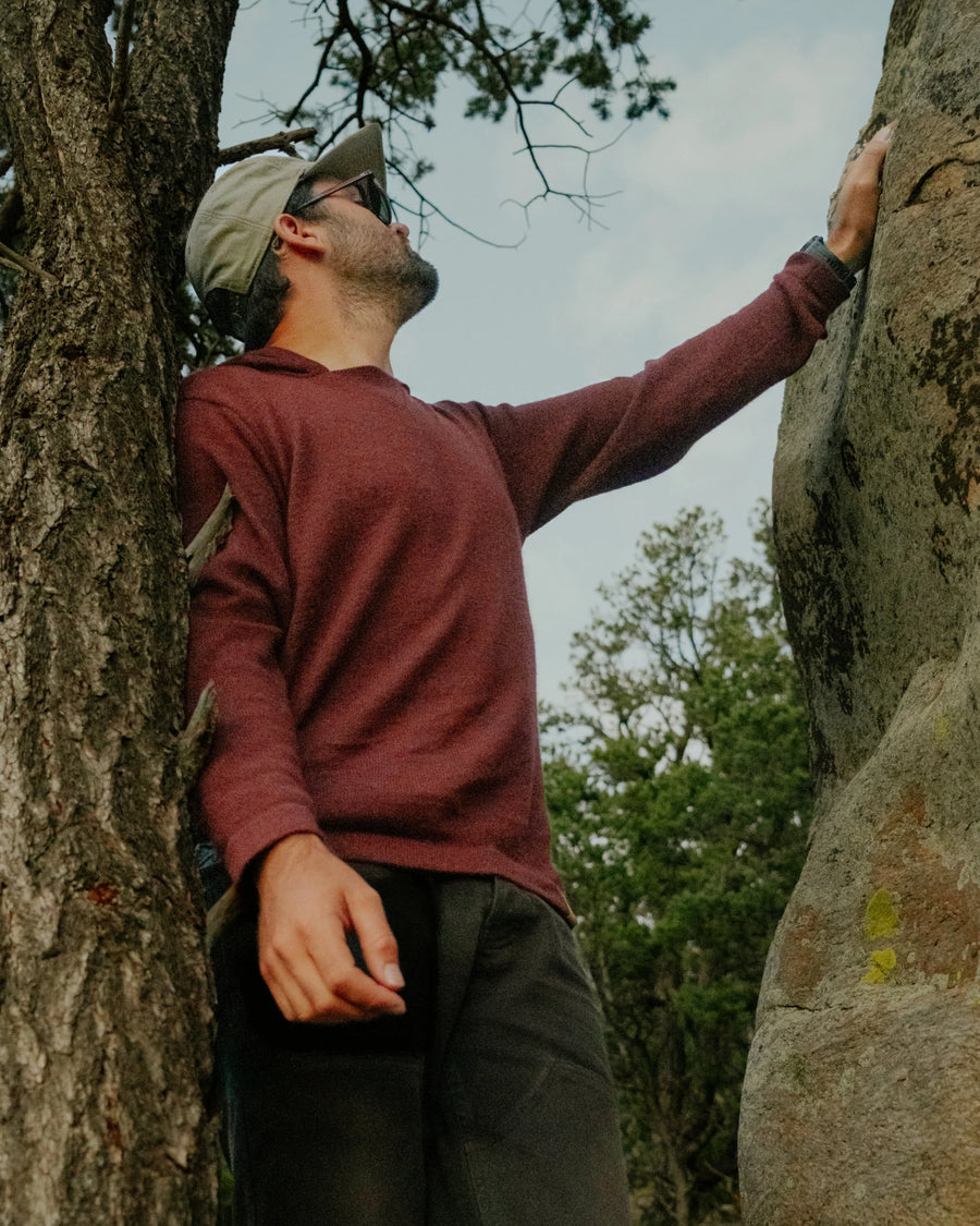 Low-angle view of burgundy alpaca hoodie on model showcasing textured knit, hood, and relaxed fit with trees and rocks background