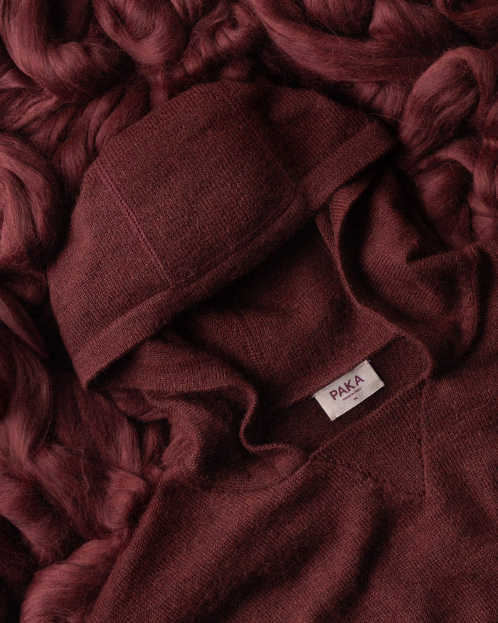 Flat lay of burgundy alpaca hoodie showing hood, soft knit texture and PAKA label on matching fiber background