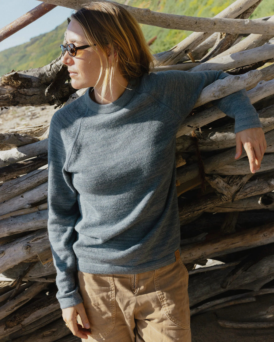 Woman wearing bluestone alpaca wool crewneck sweater in a relaxed fit leaning against driftwood on a sunny beach