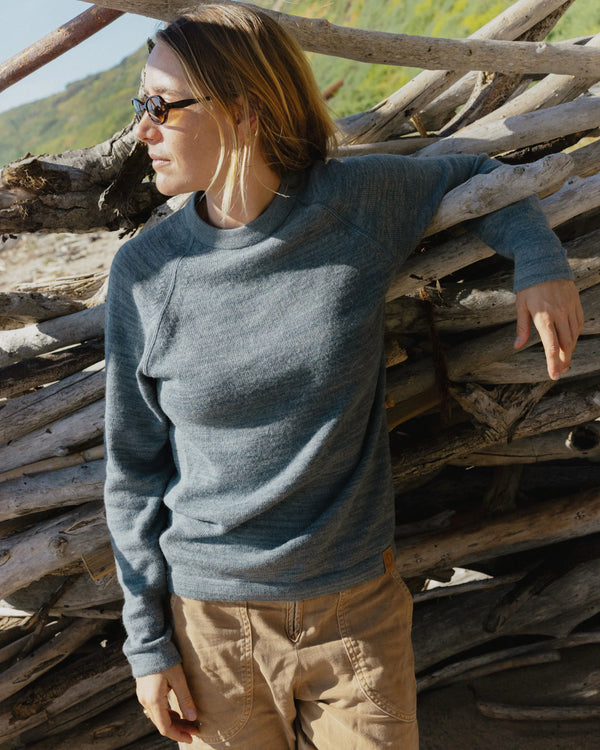 Woman wearing bluestone alpaca wool crewneck sweater in a relaxed fit leaning against driftwood on a sunny beach