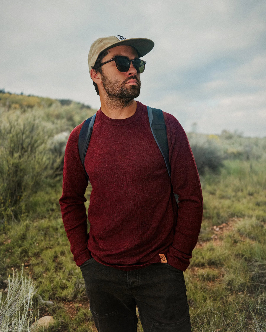 Man wearing PAKA's Original Crew in Burgundy and sunglasses with a backpack, standing in a natural landscape.