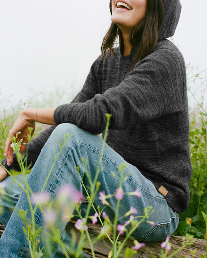 Seated three-quarter view of The Hoodie alpaca hoodie showcasing hood and textured charcoal alpaca knit with a grassy wildflower background