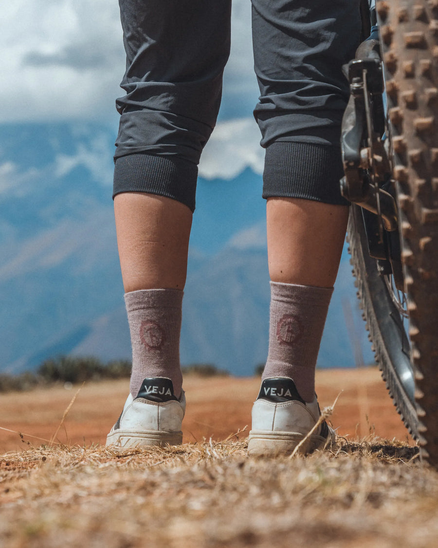 Rear view of person standing on dirt trail wearing taupe Trail 3/4 crew hiking socks and white sneakers beside mountain bike
