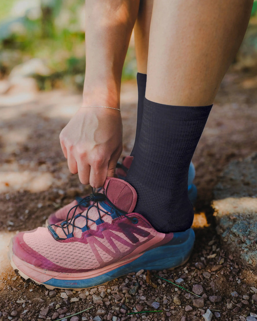 Close-up of black Trail 3/4 crew sock showing mid-calf fit and ribbing with model tying pink trail shoe on dirt path