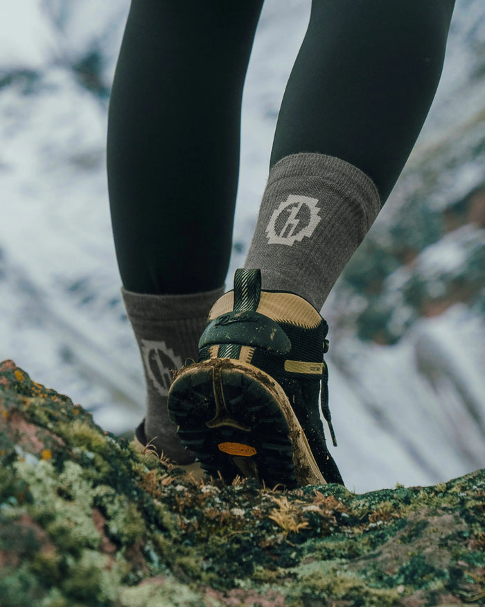 Rear view of Trail 3/4 crew sock showcasing cushioned ankle support and PAKA logo with muddy hiking shoe on a rocky trail