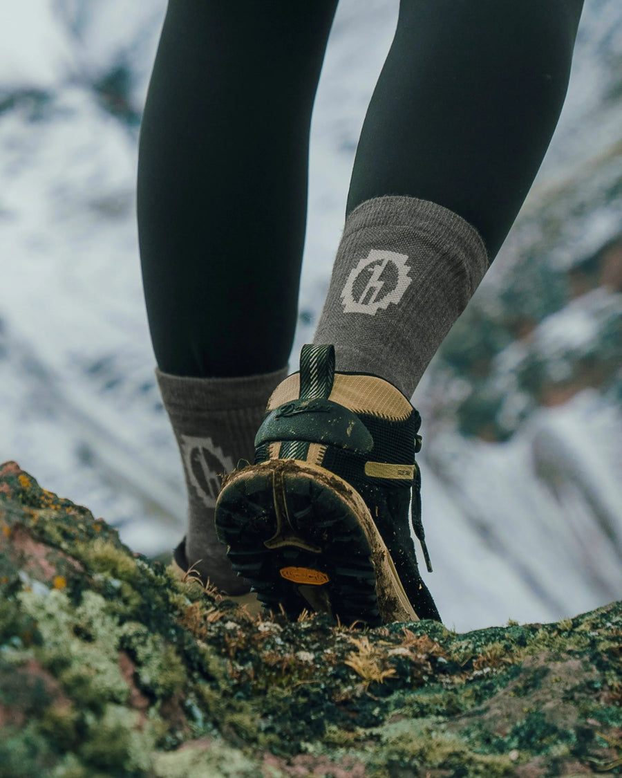 Rear view of Trail 3/4 crew sock showcasing cushioned ankle support and PAKA logo with muddy hiking shoe on a rocky trail