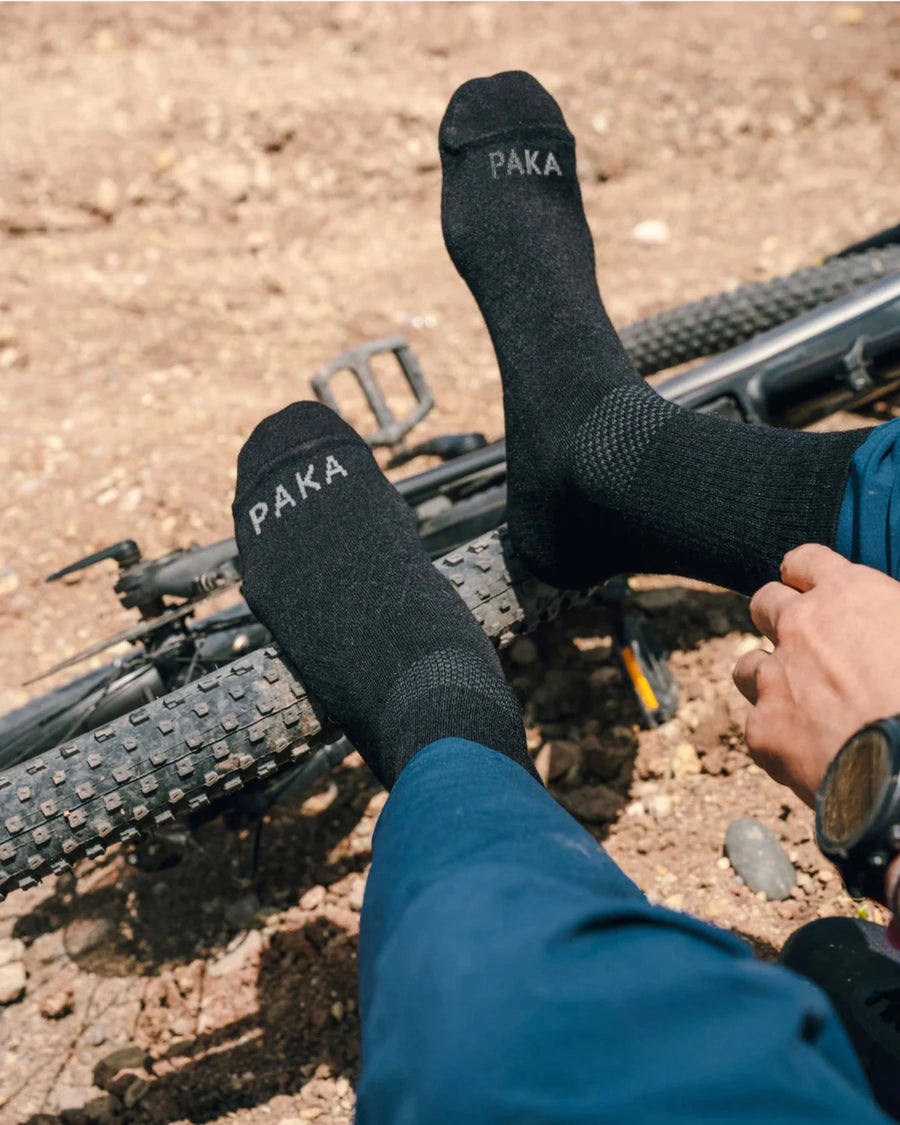 Person wearing black Trail 3/4 crew socks with PAKA on the toes, feet resting on a mountain bike tire on a rocky trail