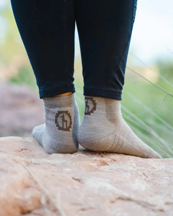 Rear view of charcoal blue Trail 1/4 Crew socks on a model showing low-crew height, heel logo, and cushioned heel on rocky outdoor background