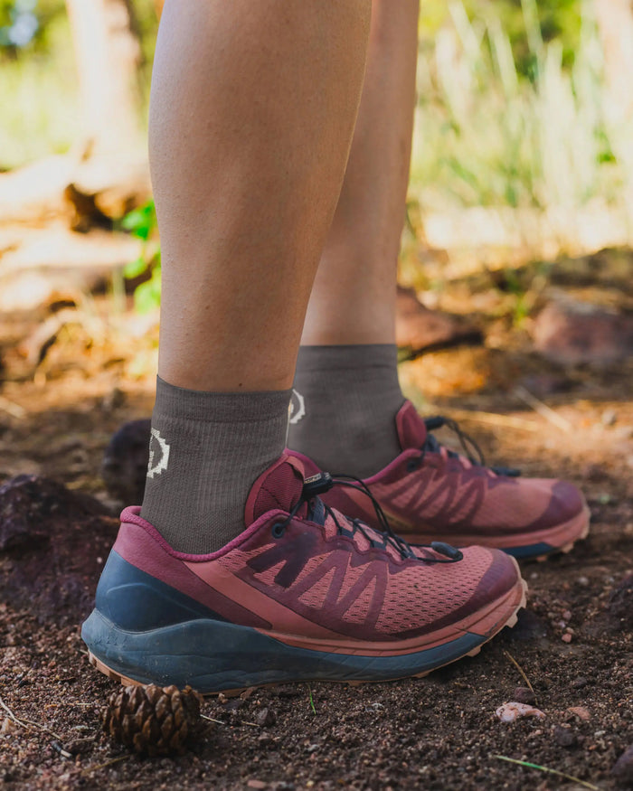 Side view of Trail 1/4 Crew lightweight hiking socks in charcoal with white logo, worn on model on a rocky trail