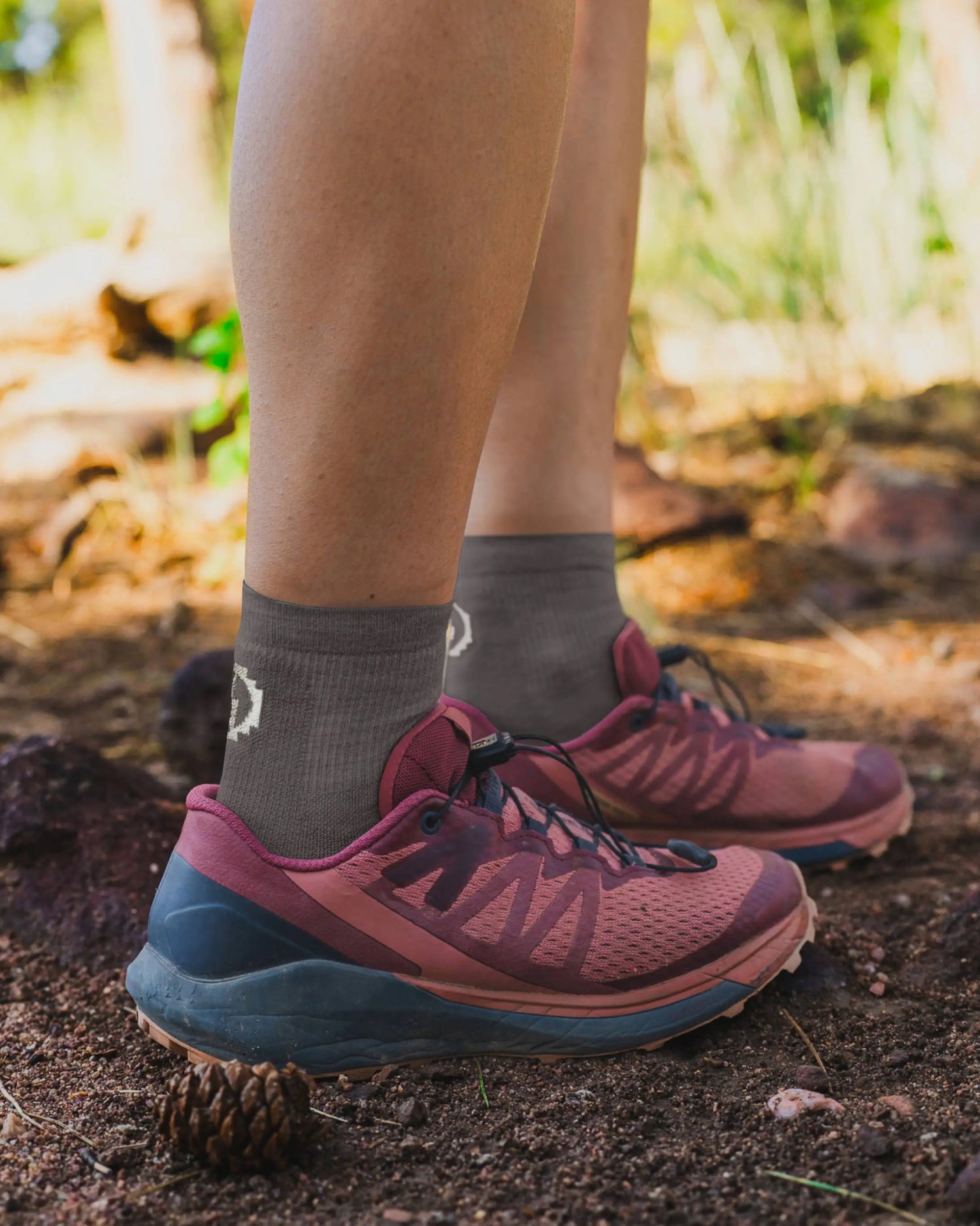 Side view of Trail 1/4 Crew lightweight hiking socks in charcoal with white logo, worn on model on a rocky trail