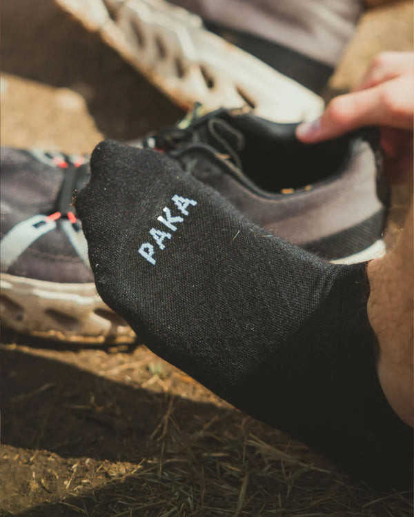 Runner pulling on black ankle alpaca socks with PAKA logo beside a trail running shoe on a dirt path