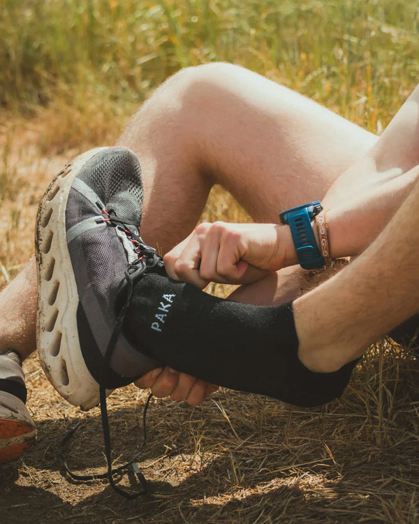 Side view of black Trail Ankle Socks on a runner’s foot showing PAKA logo and low-cut ankle fit on grassy trail