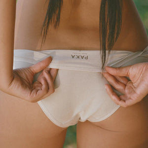 Rear view of women’s beige high-rise briefs showcasing PAKA logo on folded waistband with blurred outdoor background