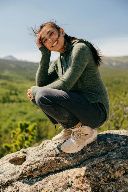 Woman crouching on a rock wearing an olive green women’s hoodie with a feminine fit in a mountain meadow