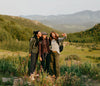 Three women standing on a grassy hillside wearing PAKA women’s hoodies with mountains in the background