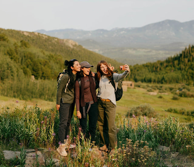 Three women standing on a grassy hillside wearing PAKA women’s hoodies with mountains in the background