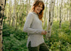 Side view of women’s light gray hoodie showing hood, drawstrings, ribbed side panels and side slit worn in a birch forest