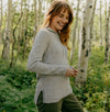 Woman smiling in a light gray women’s hoodie with ribbed side panels and relaxed feminine fit, standing in a sunlit birch forest
