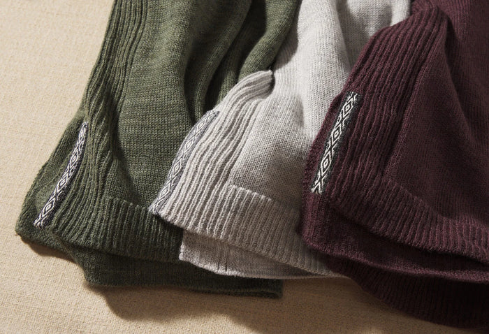 Close-up of three folded women’s hoodies (olive, light gray, burgundy) showing ribbed hems, knit texture, and woven logo tag