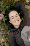 Woman lying on grass among wildflowers wearing a plum women’s hoodie, smiling with a cap resting on her chest, relaxed outdoor setting