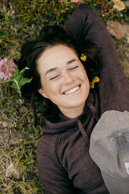 Woman lying on grass among wildflowers wearing a plum women’s hoodie, smiling with a cap resting on her chest, relaxed outdoor setting