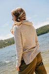Rear three-quarter view of women’s tan hoodie showing hood and relaxed fit on a woman standing by a lake and hills