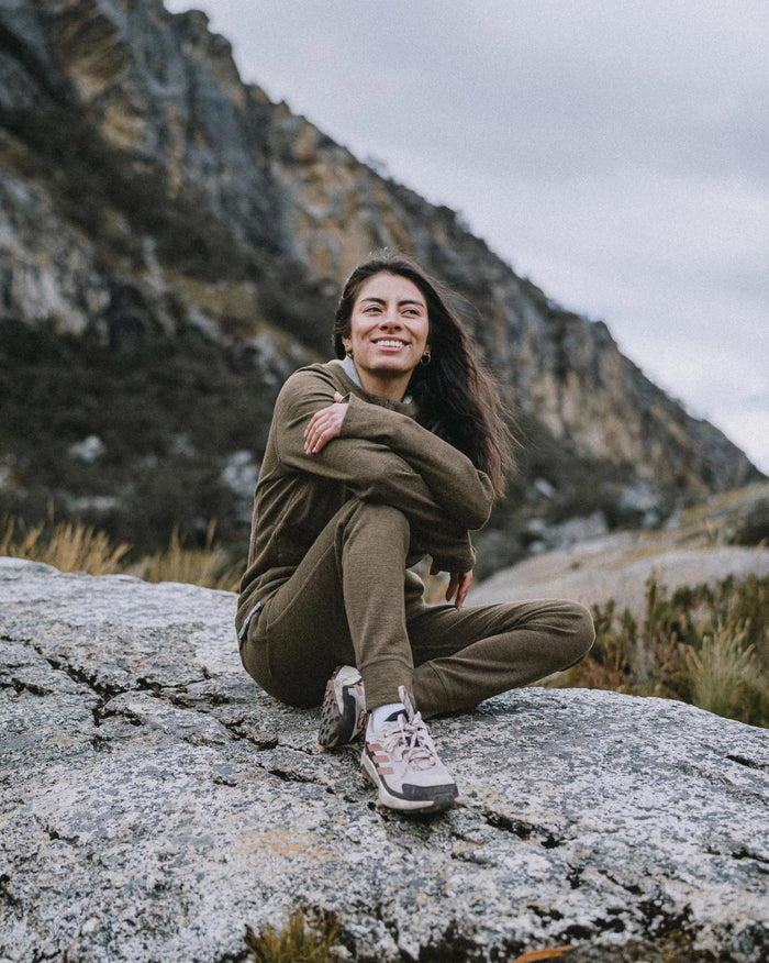 Woman sitting on a rock wearing Andean Moss women’s Breathe alpaca joggers, smiling in a mountain outdoor setting