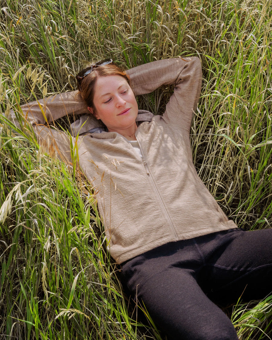 Woman lying in tall grass wearing a light beige alpaca full-zip hoodie in a relaxed outdoor setting