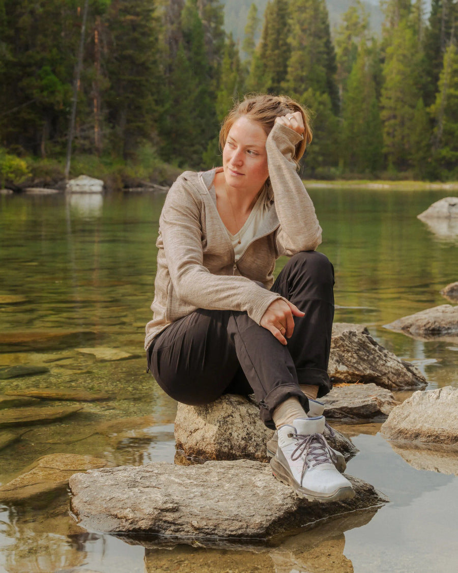 Woman wearing a beige Women’s Breathe full-zip alpaca hoodie sitting on rocks by a mountain lake, relaxed outdoor setting