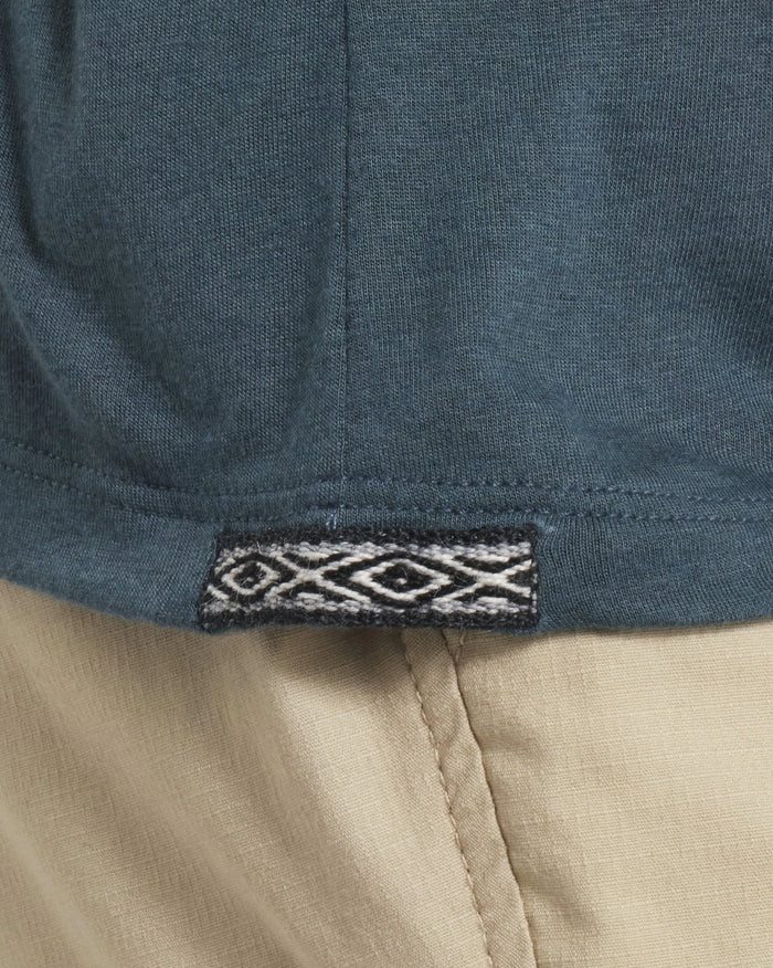 Hem close-up of Women’s Aire Long Sleeve shirt showcasing woven Inca tag on navy fabric with beige pants background